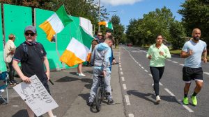 Young people take part in an anti-immigration protest outside the former Crown Paints factory in Coolock, north of Dublin, July 2024, while a couple of joggers pass by. The building has been earmarked to house asylum seekers in Dublin. Photo: Paul Faith/AFP/Getty