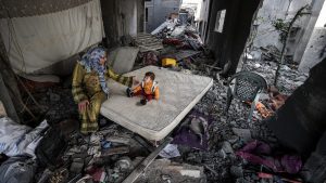 A Palestinian woman and a baby sit on a mattress among the rubble of their family home in Deir al Balah, Gaza, which was destroyed by Israeli attacks in November 2023. Photo: Ali Jadallah/Anadolu/Getty